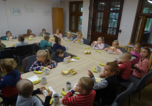 Grupa dzieci siedzi przy stołach na których przed każdym dzieckiem leży talerzyk z ciastem oraz bananem. Część dzieci trzyma w ręku ciasto i rozpoczyna jedzenie,